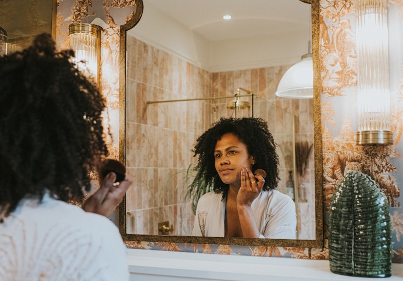 A beautiful young black woman applies foundation / concealer with a make-up brush in a bathroom mirr...