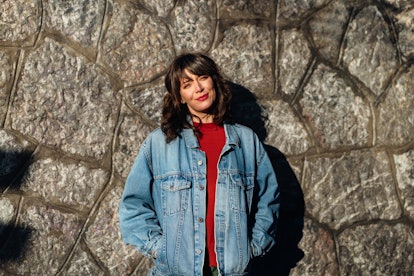 Cropped photo of a beautiful woman in denim jacket leaning against a stone wall. She is wearing a re...