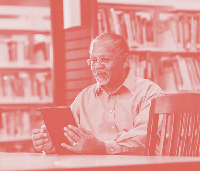 A senior African-American man in his 60s sitting at a table in a public library, using a digital tab...