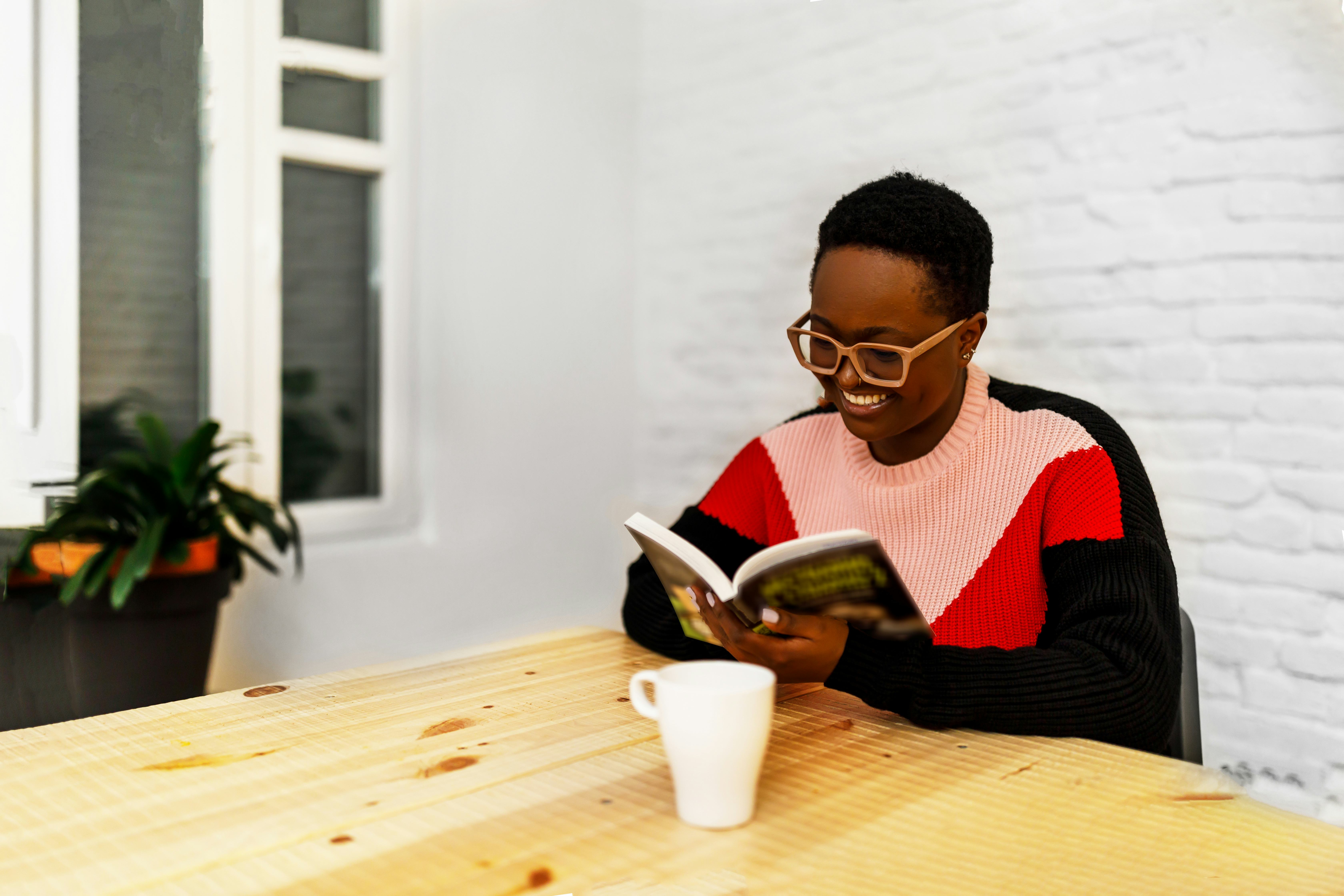 Smiling black woman sitting for her table reading a book at home in the dining room.