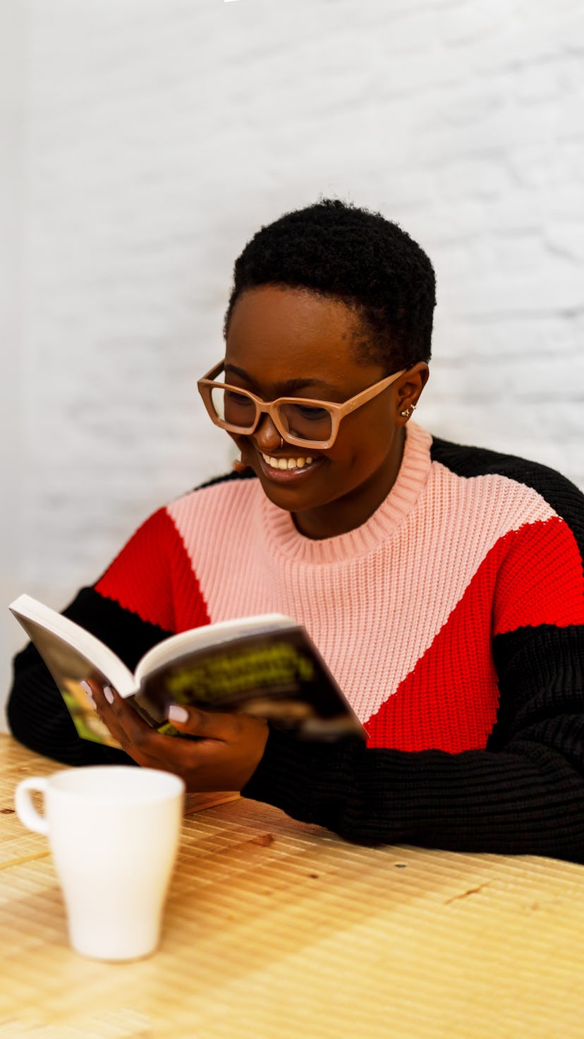 Smiling black woman sitting for her table reading a book at home in the dining room.