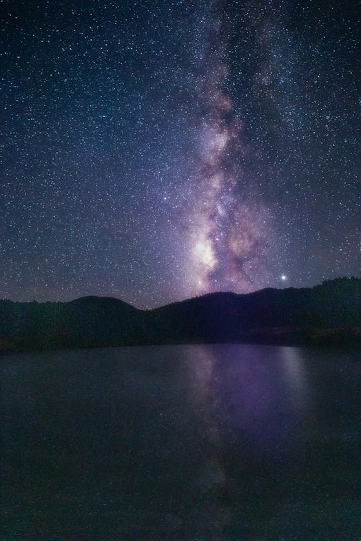 The glowing purple band of the Milky Way shines vertically over a body of water. 