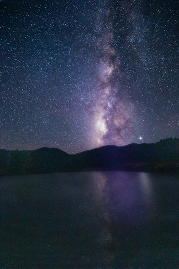 The glowing purple band of the Milky Way shines vertically over a body of water. 