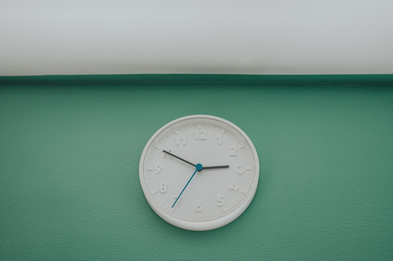 A simple white wall clock hangs above a sunny window with blinds. It displays the time, 2.49. The tu...