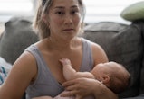 A middle aged mother sitting on her couch holds her baby in her arm with a tired and stressed expres...