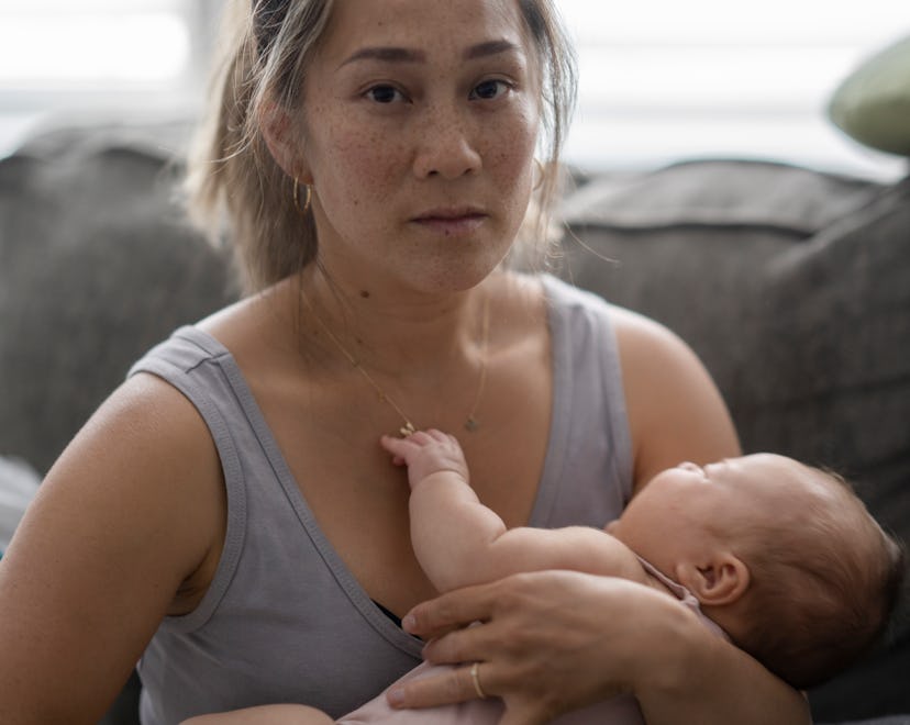 A middle aged mother sitting on her couch holds her baby in her arm with a tired and stressed expres...