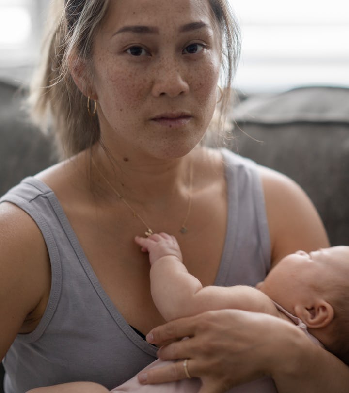 A middle aged mother sitting on her couch holds her baby in her arm with a tired and stressed expres...