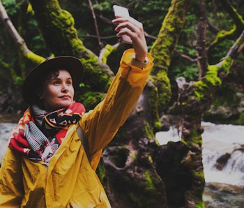 Young woman wearing yellow raincoat and backpack hiking in the forest on a rainy day, taking photos ...