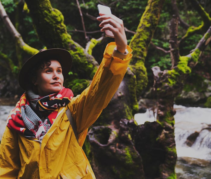 Young woman wearing yellow raincoat and backpack hiking in the forest on a rainy day, taking photos ...