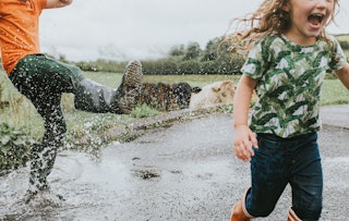 Two cute kids jump in a huge puddle, wearing welly boots. Boy playfully kicks water towards girl as ...
