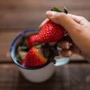 several strawberries in a white enamel mug and one hand holding a strawberry