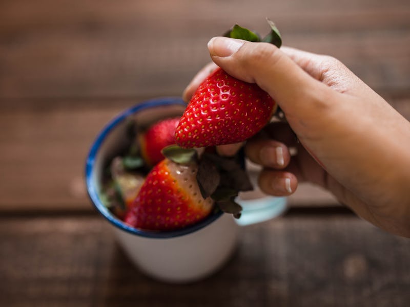 several strawberries in a white enamel mug and one hand holding a strawberry