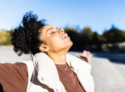 Relaxed afro woman enjoying life outdoors while standing with her arms opened and eyes closed. Freed...