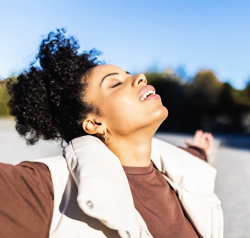 Relaxed afro woman enjoying life outdoors while standing with her arms opened and eyes closed. Freed...