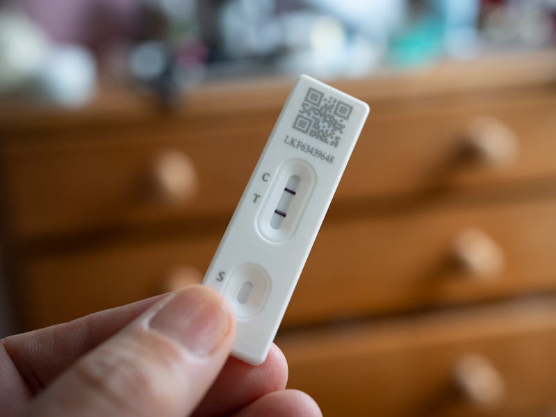 A detail of a hand holding a lateral flow test that shows a positive result in a domestic bedroom, o...