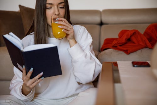 Shot of focused young woman sitting on the living room floor, drinking orange juice and reading an i…