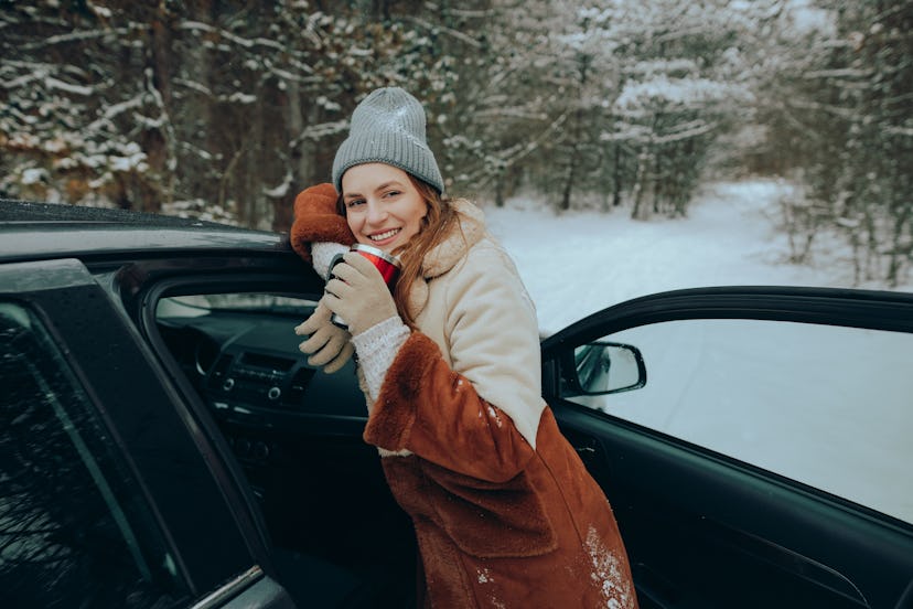Smiling young woman wearing a knit hat leaning on the car and holding a travel mug filled with a hot...