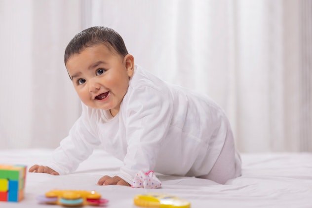 Brown haired Indian baby boy dressed in white smiles at camera as he crawls.