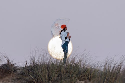 A woman stands with a clear umbrella in front of the full moon. These may 2022 full flower moon rit...