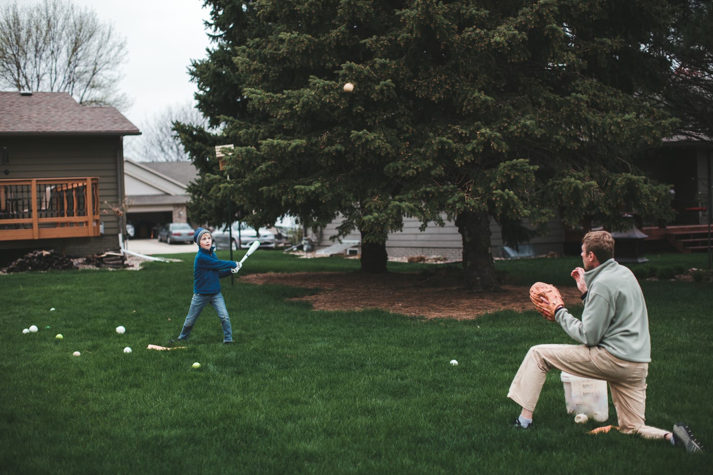 Boy hits a baseball into the air with a bat that his father has just pitched to him while practicing baseball in their backyard.