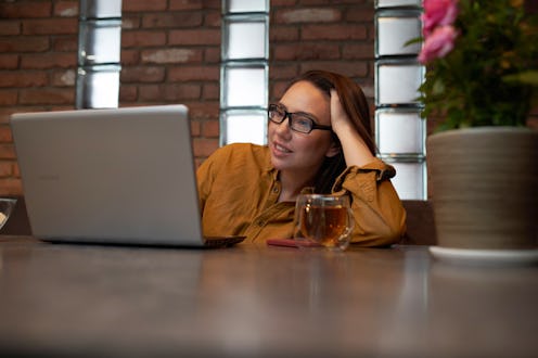 Positive female in glasses touching head and reading data from laptop with smile while sitting at ta...