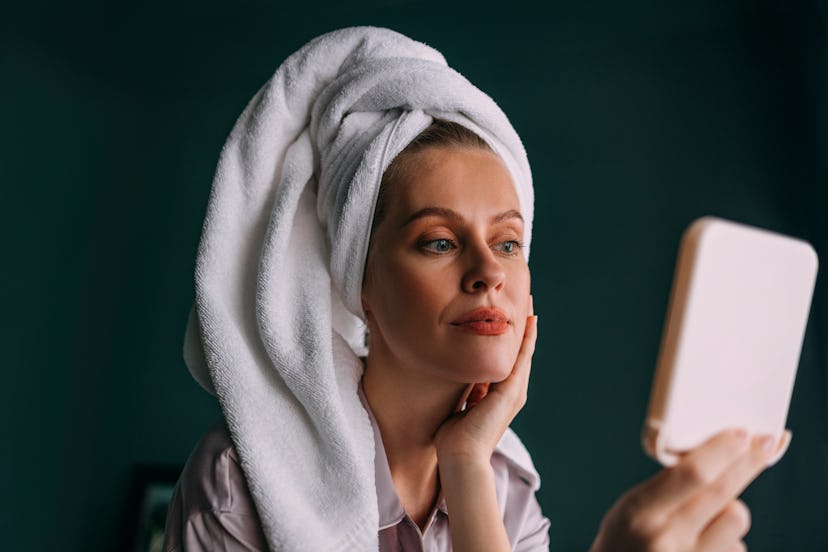 A charming Caucasian woman with her hair wrapped up in a towel smiling while applying some creme on ...