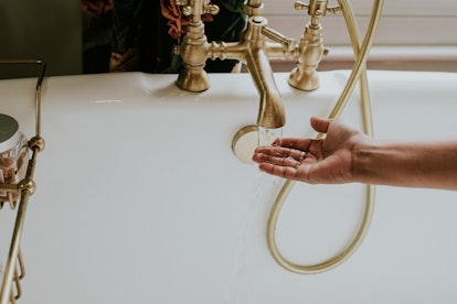 A black woman holds her hands under the gold faucet of a roll top bath to access the temperature of ...