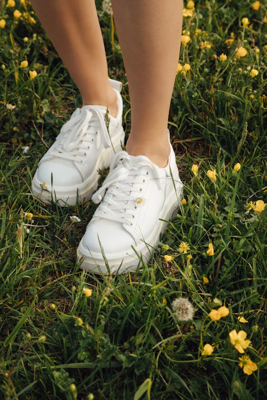 legs of a young European woman in white sneakers, a chamomile is inserted into the laces in a field ...