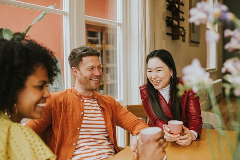 Three beautiful young people socialise at a table while enjoying hot drinks. They are animated as t...