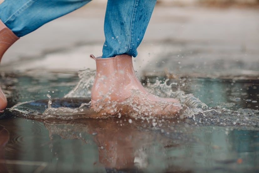Woman wearing rain rubber boots walking running and jumping into puddle with water splash and drops ...