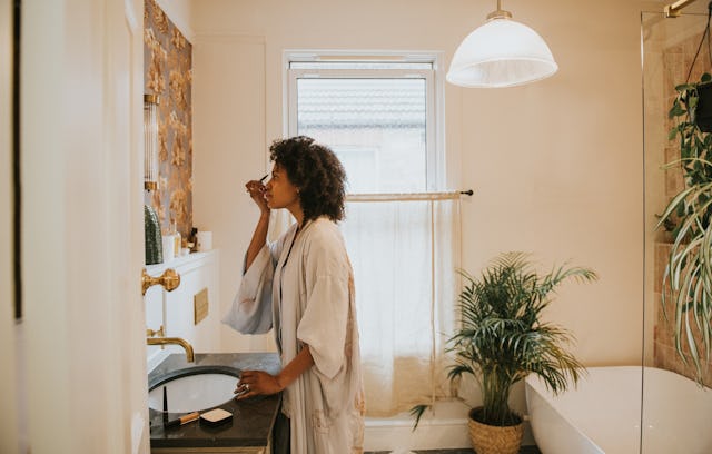 A beautiful black woman stands in a stylish bathroom in a long, silky robe and carefully applies her...