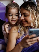 Young mother and little daughter in purple costumes using mobile phone at home