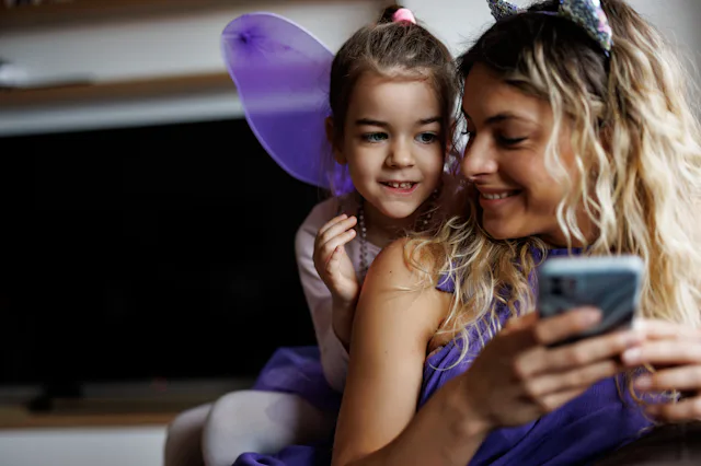Young mother and little daughter in purple costumes using mobile phone at home