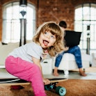 A toddler playing on a skateboard indoors while her mother works on a laptop.