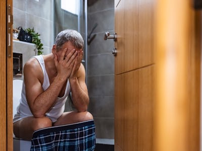 Photo of mature man on toilet is crouching forward in pain because of his stomach cramps.