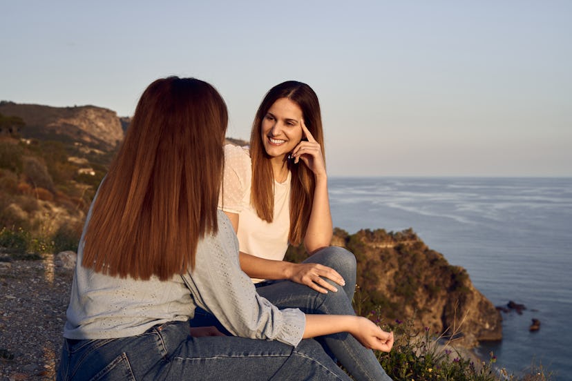 Couple of friends talking and laughing while watching the sunset from a cliff by the sea. Speak your...