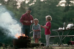 A father watches his daughters cook marshmallows over the campfire.