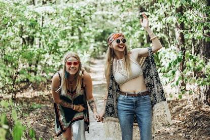 Two nickering young hippie women walking in summer forest holding hands after reading their June 202...
