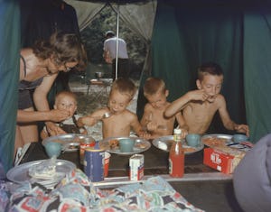Under a tent that extends over their station wagon, the Baldwin family eats a meal from metal plates...