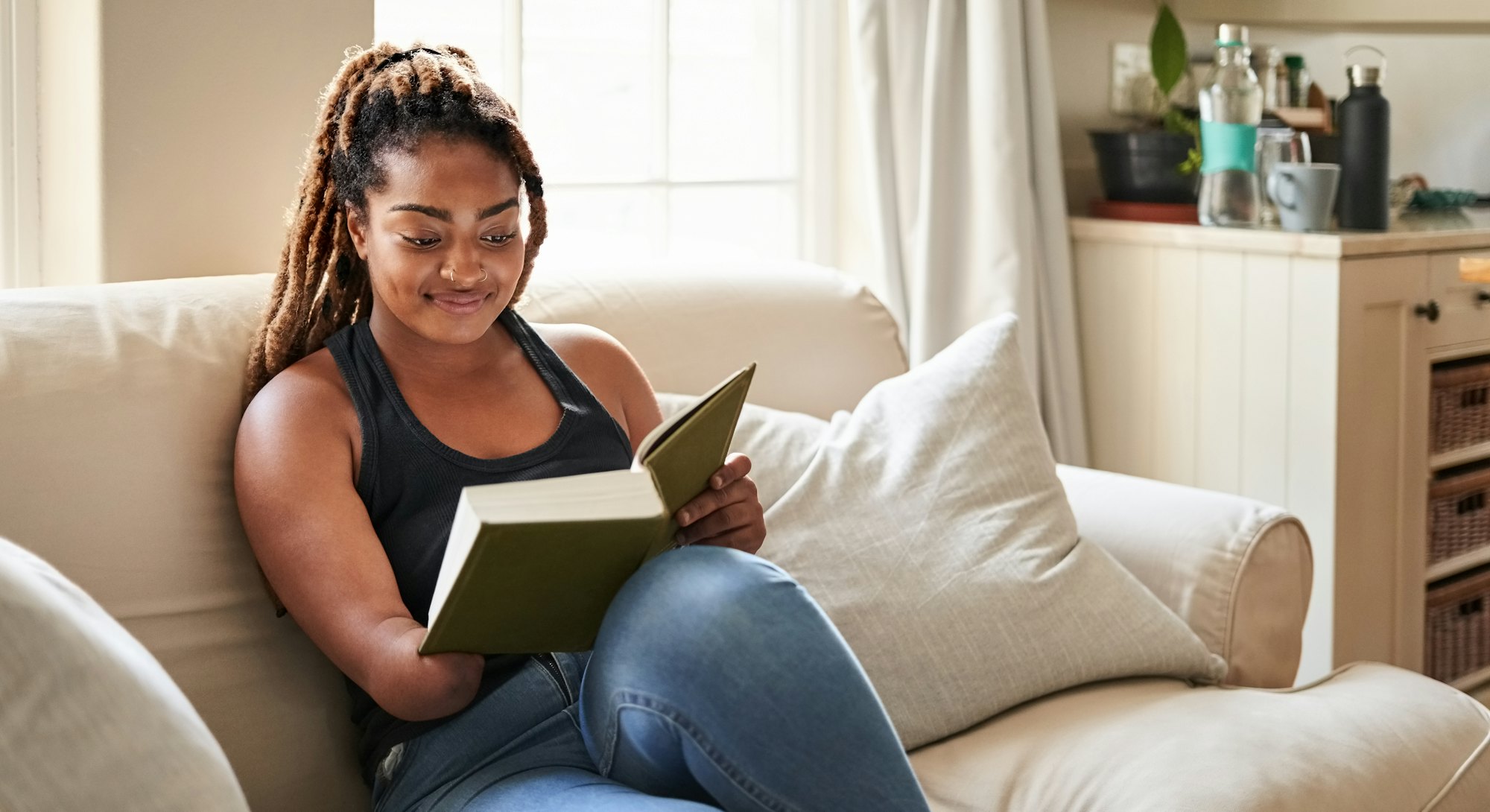 Young African woman reading a book while sitting on living room sofa at home, woman missing lower ha...