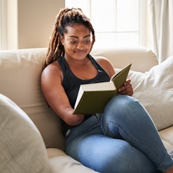 Young African woman reading a book while sitting on living room sofa at home, woman missing lower ha...