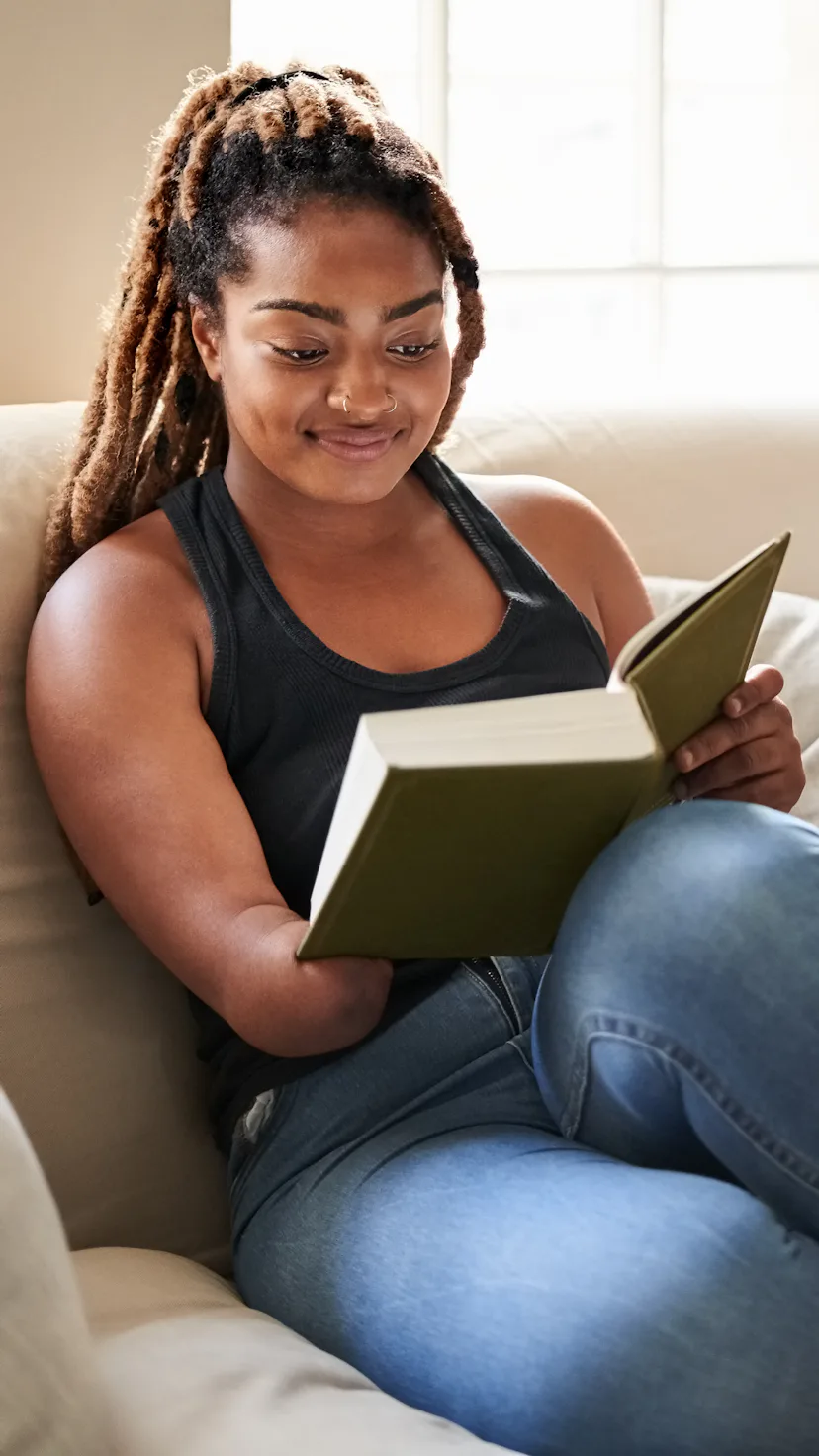 Young African woman reading a book while sitting on living room sofa at home, woman missing lower ha...