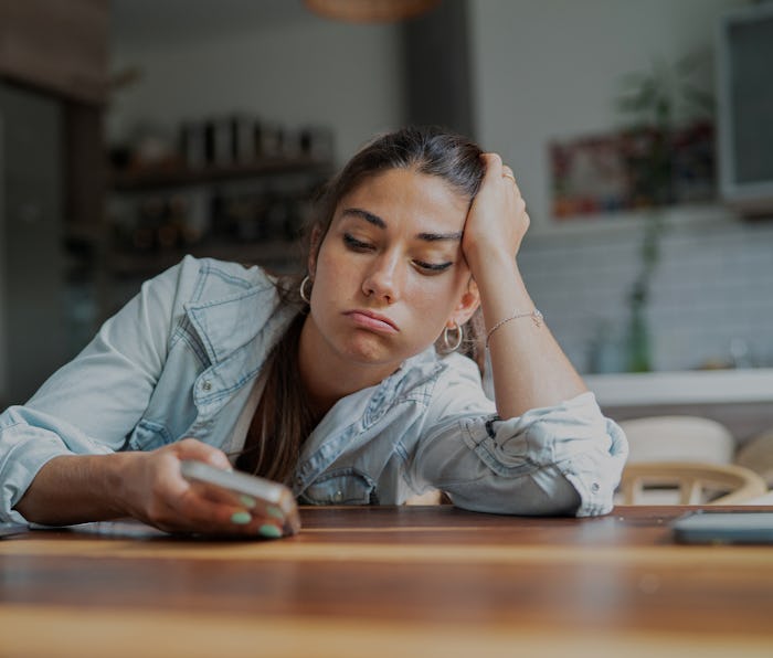 Portrait of bored young adult woman looking at smart phone while leaning on hand during daytime