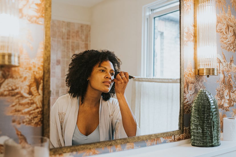 A beautiful black woman stands in a stylish bathroom in a long, silky robe and carefully applies her...