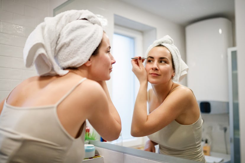 Reflection of woman in bathroom mirror wearing towel on wet hair, tweezing her eyebrow