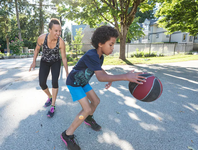A boy is dribbling a basketball while his mom marks him from behind