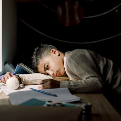 A boy with ADHD lays his head on his desk and plays with a ball while doing homework.