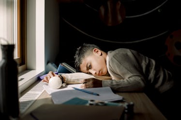 A boy with ADHD lays his head on his desk and plays with a ball while doing homework.