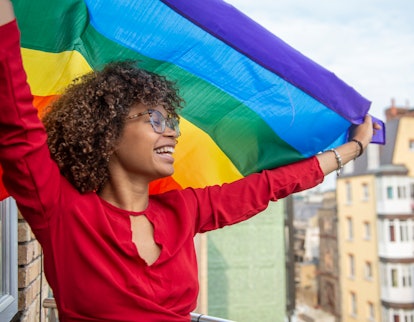 A young lady waving a gay pride flag in support of Gay Pride on her balcony needs Pride Month 2022 c...