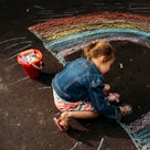 A child draws a rainbow on asphalt with chalk.